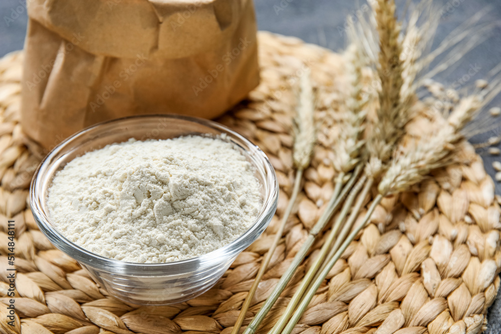 Bowl with flour and wheat ears on wicker mat, closeup