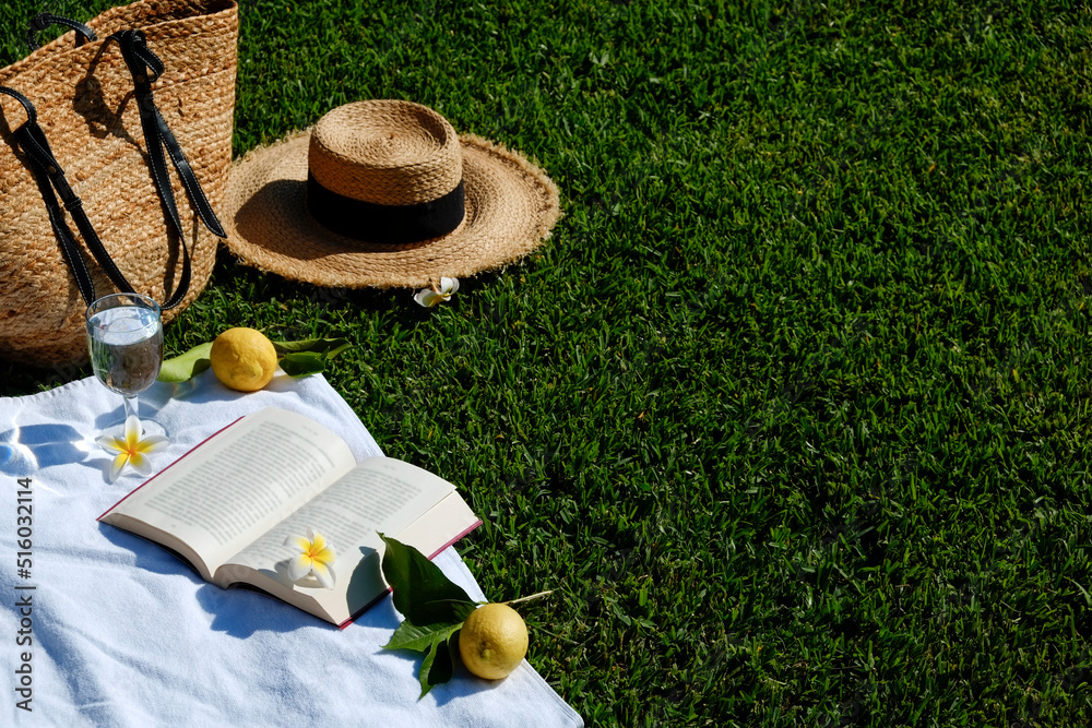 Lunch break in the park. Picnic blanket with an open book, lemons, beach bag and broad brim straw ha