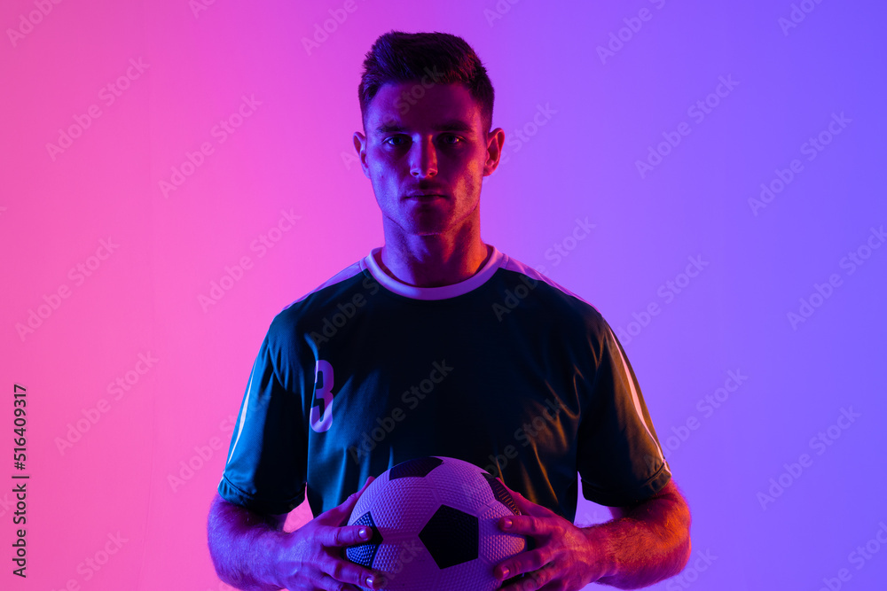 Portrait of caucasian male soccer player with football over neon pink lighting