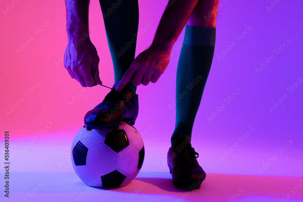 Caucasian male soccer player with football tying shoes over neon pink lighting