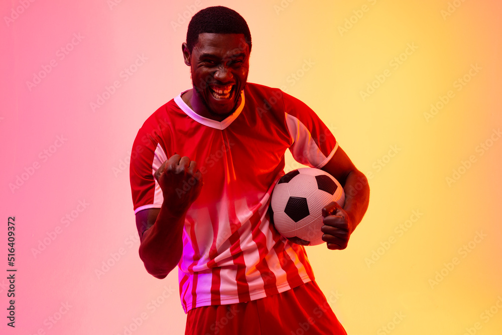 Portrait of happy african american male soccer player with football over neon pink lighting