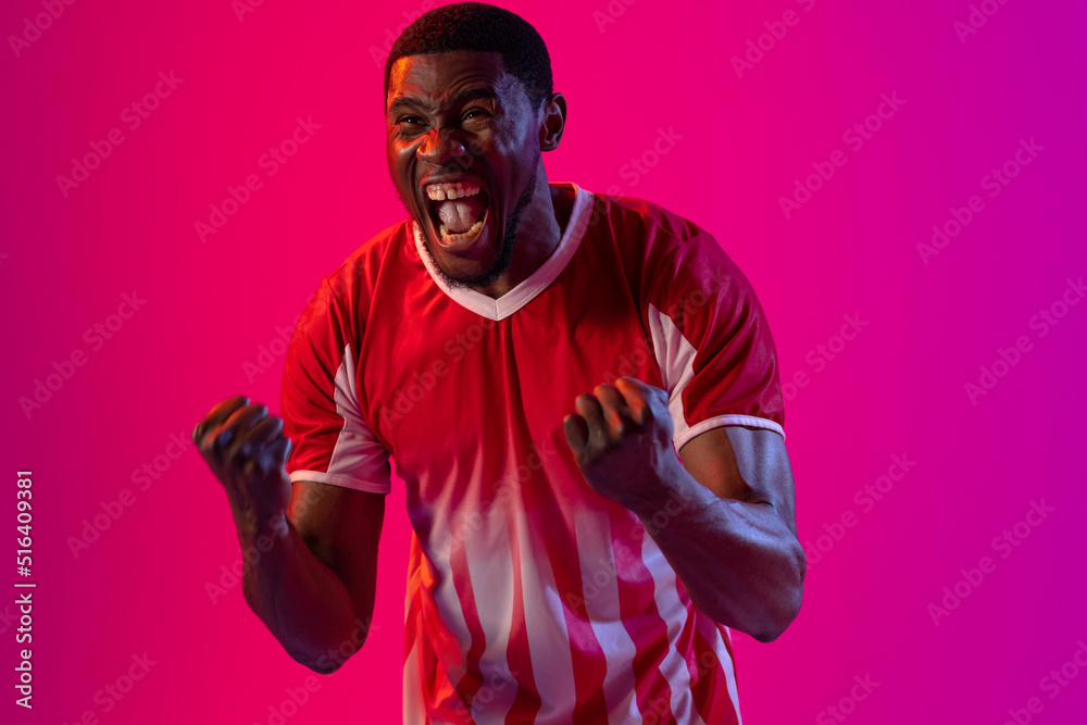 Happy african american male soccer player raising hands over neon pink lighting