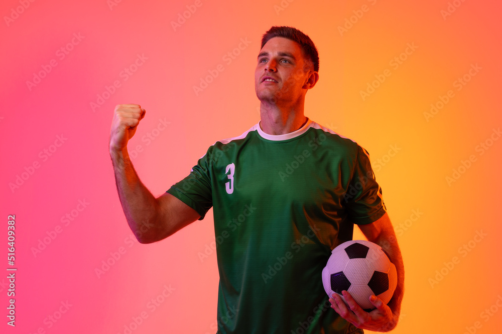 Caucasian male soccer player raising hand with football over neon pink lighting