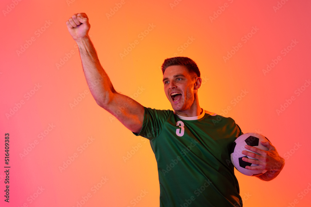 Happy caucasian male soccer player raising hand with football over neon pink lighting