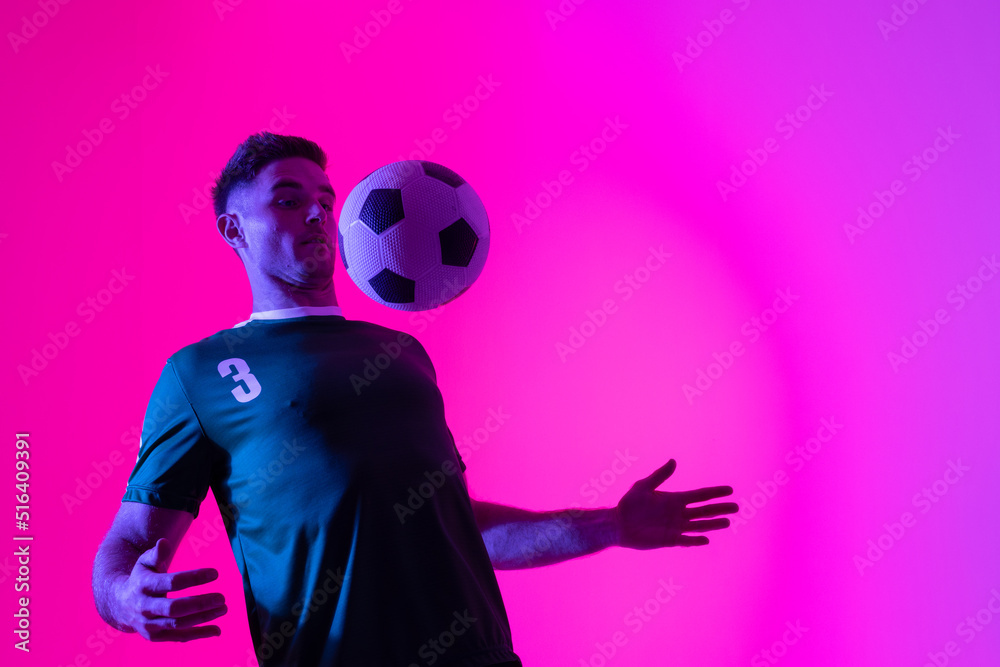 Caucasian male soccer player playing with football over neon pink lighting