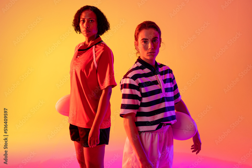 Portrait of diverse female rugby players with rugby balls over neon pink lighting