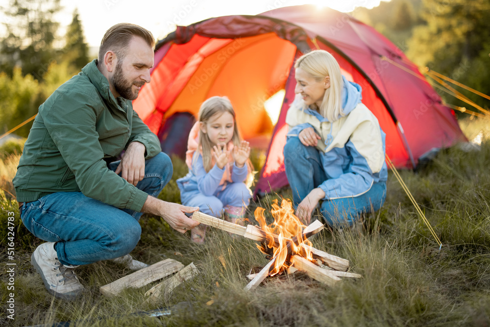 Young adult couple sit with a little girl by the campfire at campsite, traveling with tent on nature