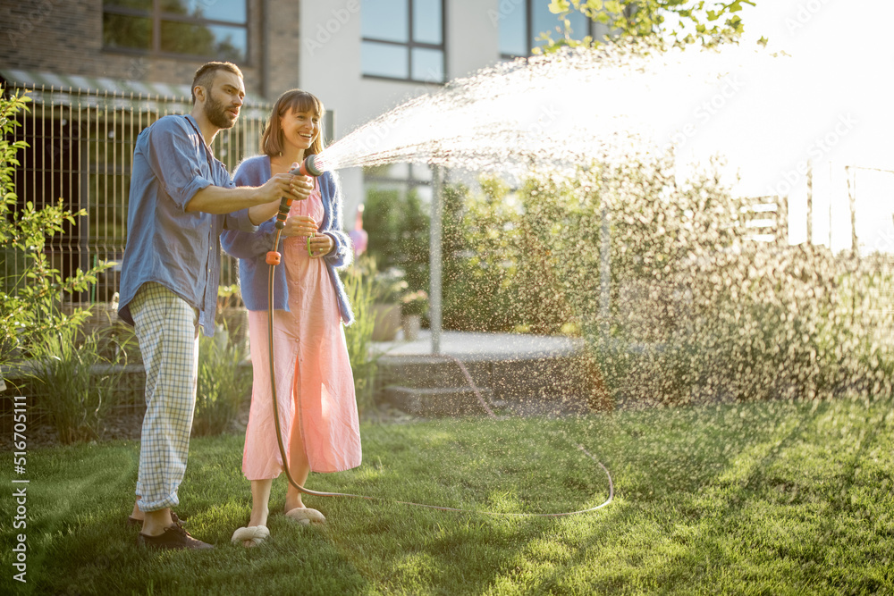 Lovely couple watering lawn at backyard of their country house on sunset. Young family taking care o