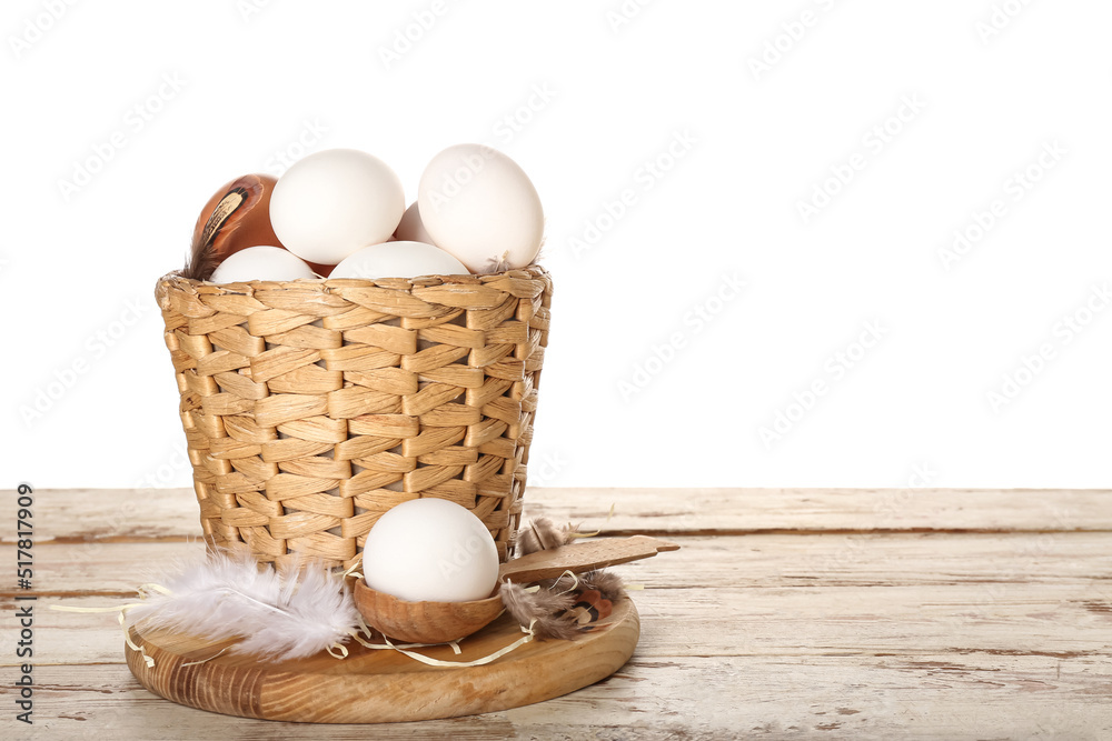 Board with wicker basket of chicken eggs and feathers on wooden background against white background