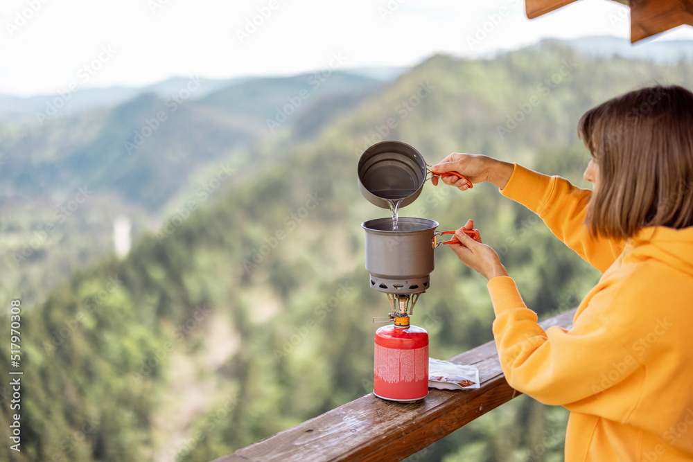 Young woman prepares food in special dishware for hiking and portable gas burner, while traveling in