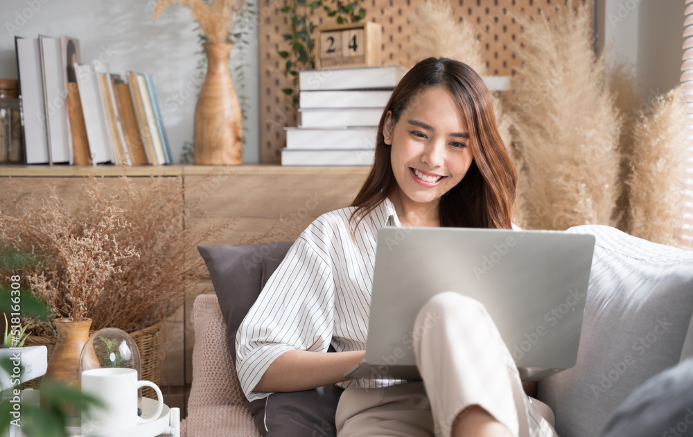 Young Asian woman using laptop on sofa living room at home cozy happy working at home.