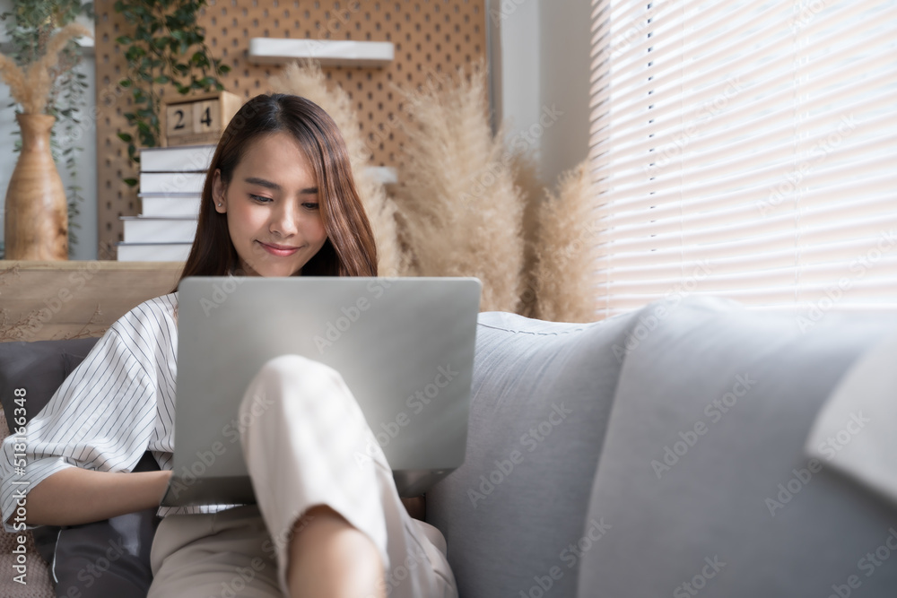 Young Asian woman using laptop on sofa living room at home cozy happy working at home.