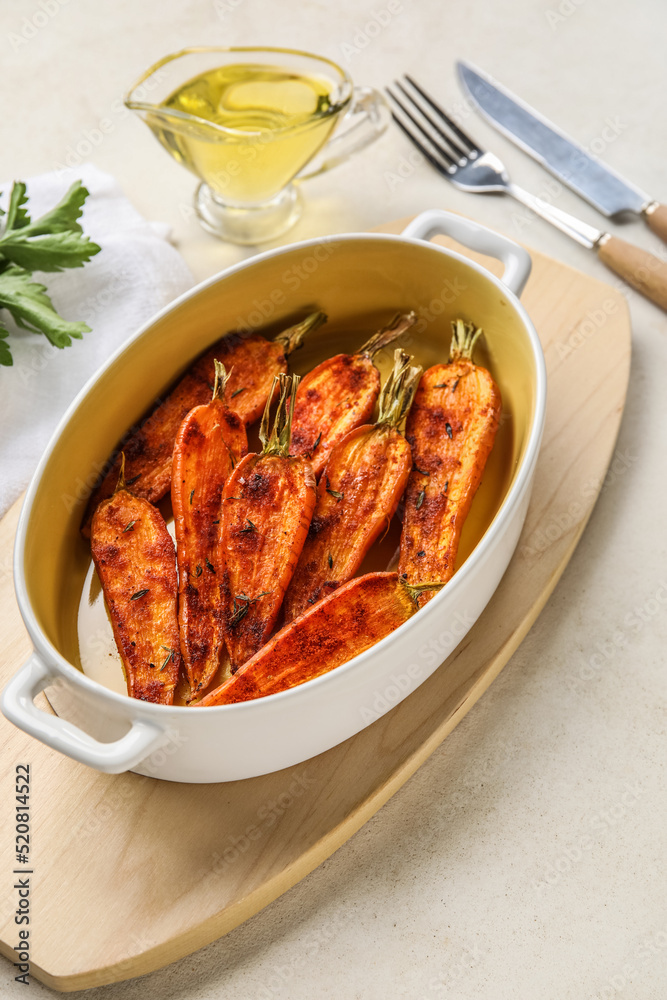 Wooden board with baking dish of tasty baked carrots on light background, closeup