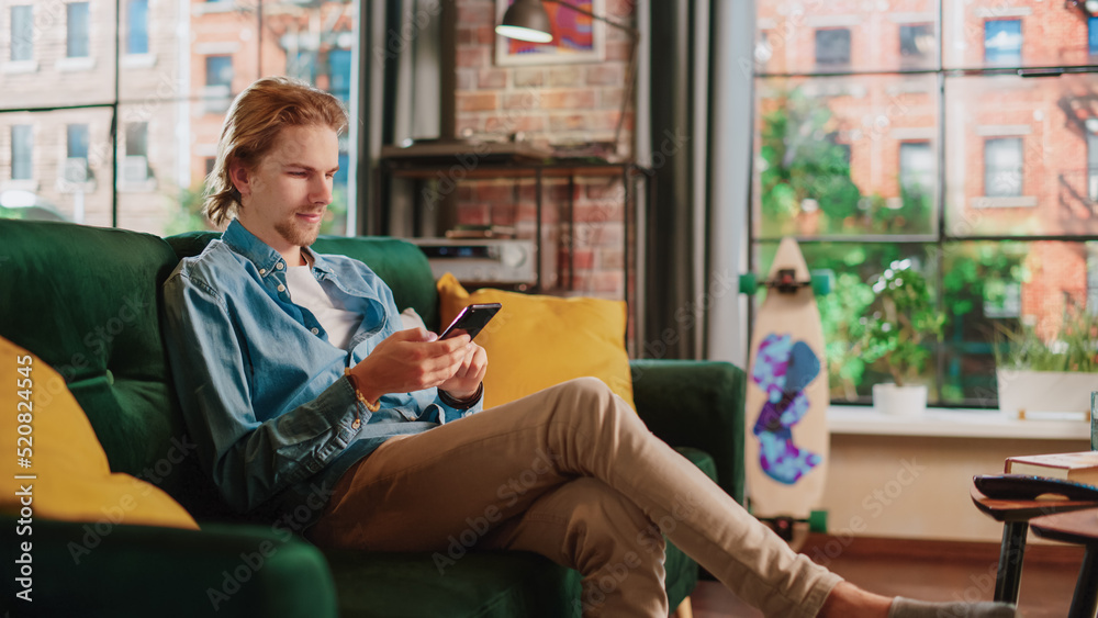 Young Handsome Adult Man Sitting on a Couch in Living Room, Relaxing and Using Smartphone. Creative 