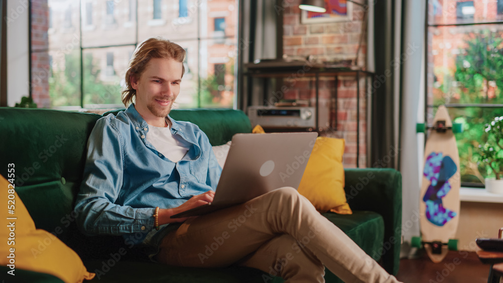 Young Handsome Adult Man Sitting on a Couch in Loft Apartment, Working from Home on Laptop Computer.