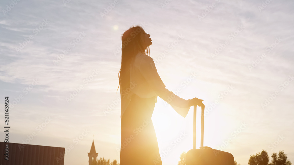 Silhouette of woman with long dark hair in business clothes stands holding suitcase at back sunset. 