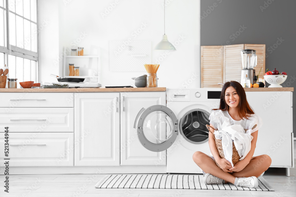 Young Asian woman with laundry basket sitting near washing machine in kitchen