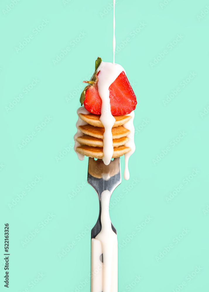 Fork with mini pancakes, fresh strawberry and pouring sauce on green background