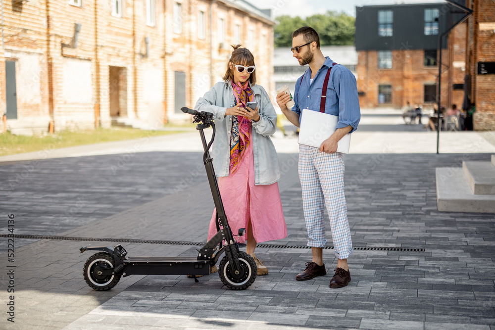 Young stylish colleagues meet together near office outdoors. Woman drives electric scooter with gree