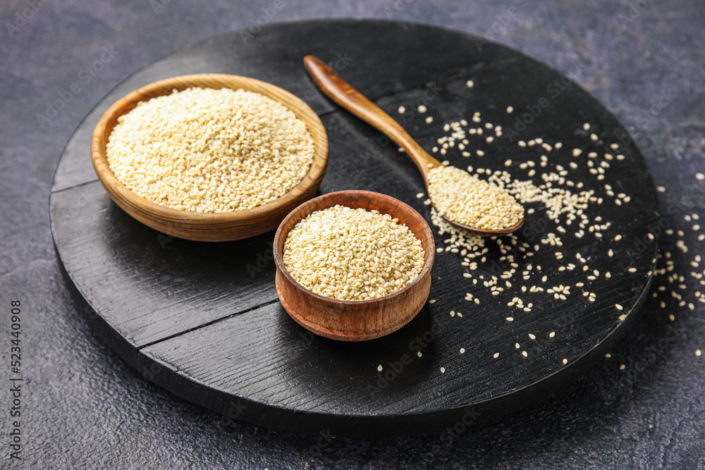 Wooden board with bowls and spoon of sesame seeds on dark background, closeup