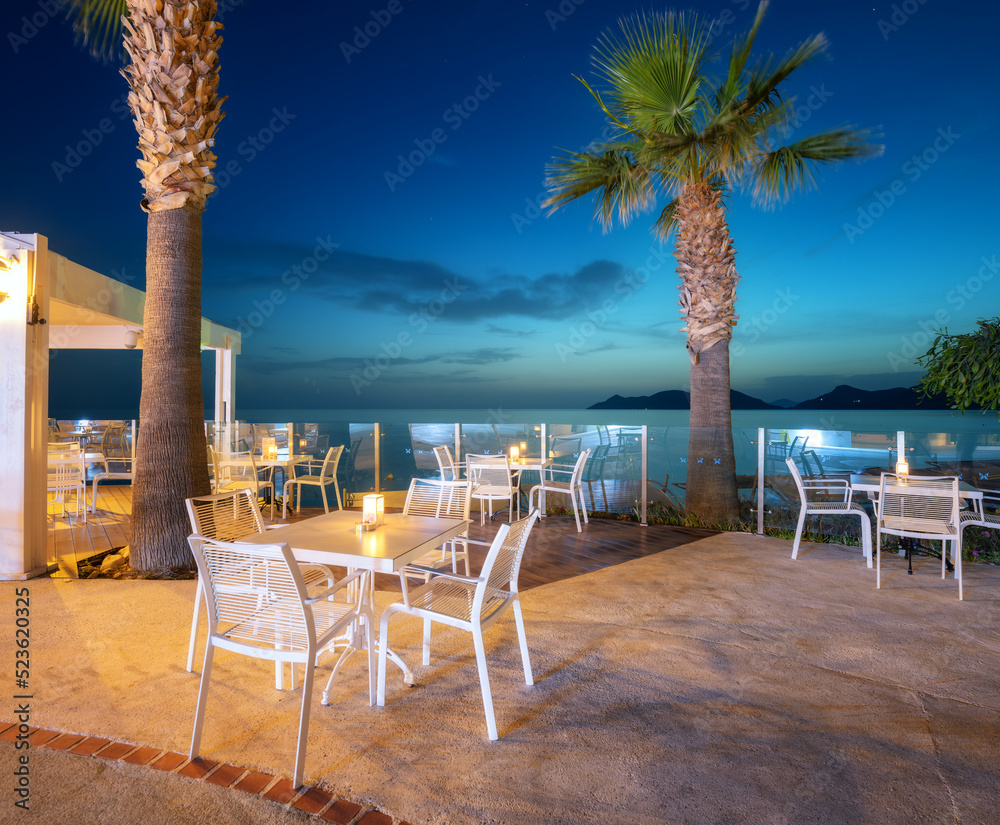 Beautiful cafe on sea coast at night in summer. Landscape with chairs and tables, lamps, lights, pal