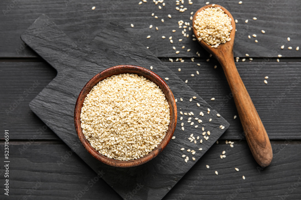 Board with bowl and spoon of sesame seeds on dark wooden background