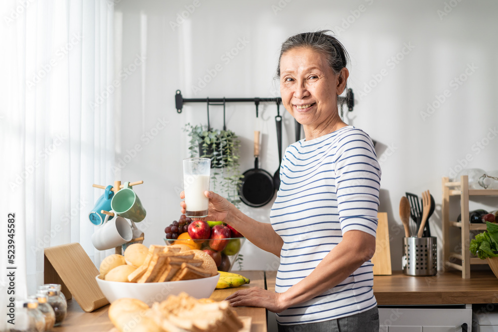 亚洲资深成熟女性在家里的厨房里喝着一杯牛奶。