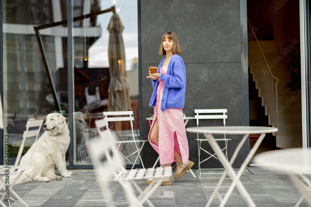 Young stylish woman goes with coffee cup from a coffee shop, cute white dog waits for her near the t