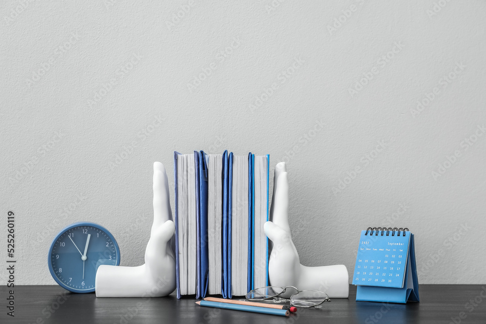 Stylish holder with books, eyeglasses, calendar and alarm clock on table near light wall