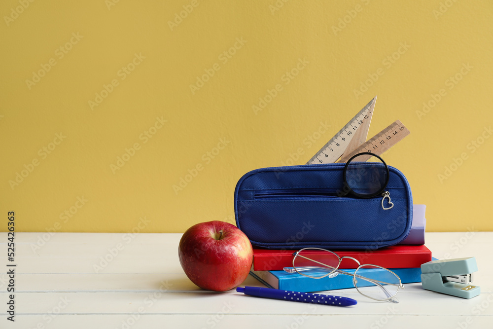 Pencil case with school stationery, eyeglasses and apple on table against yellow background