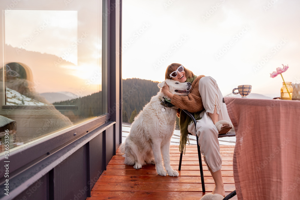 Woman hugs her dog while resting on terrace of tiny house in the mountains, enjoying beautiful lands