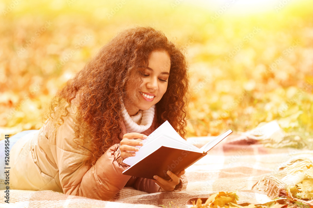 Young African-American woman reading interesting book on plaid in autumn park