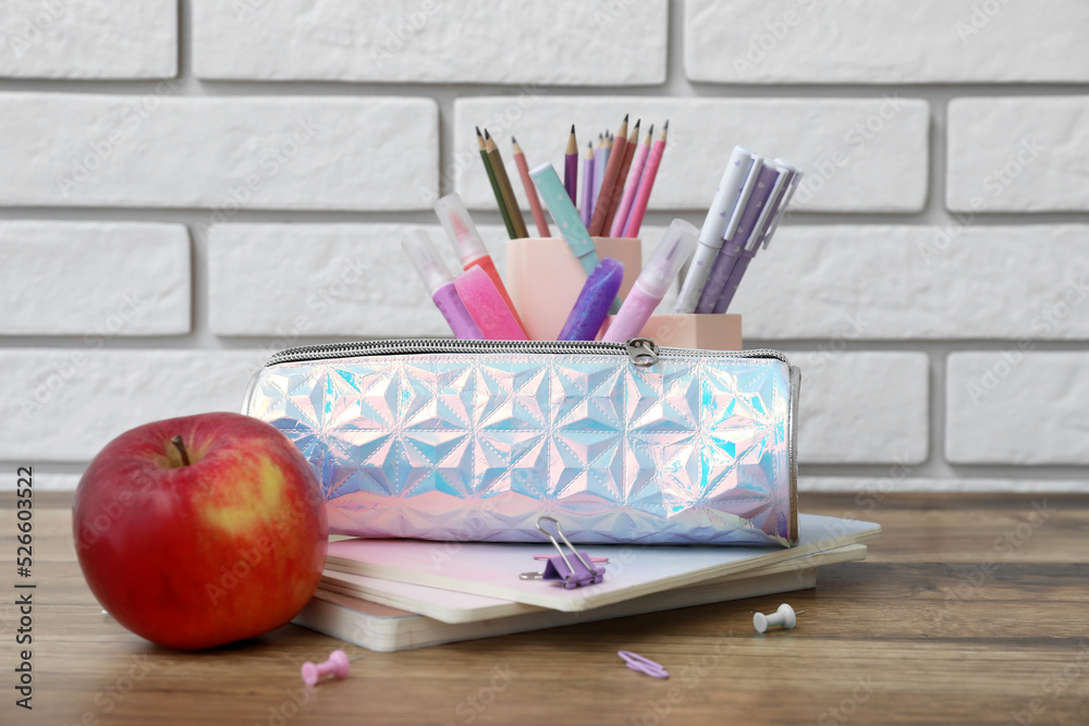 Holographic pencil case with school stationery and apple on table against white brick background