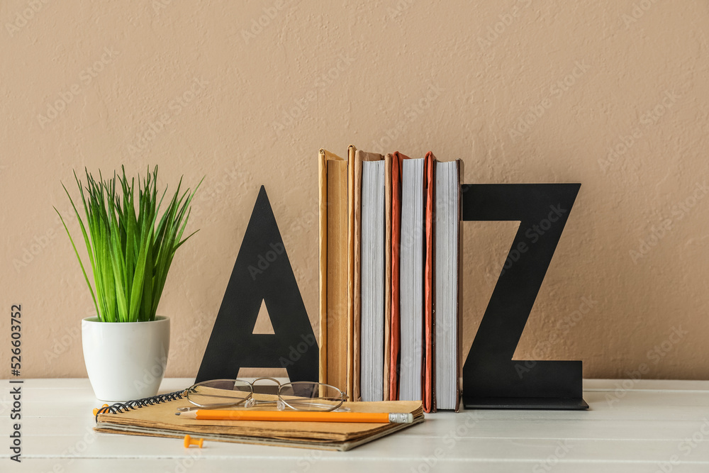 Stylish holder with books, eyeglasses and flowerpot on table near beige wall