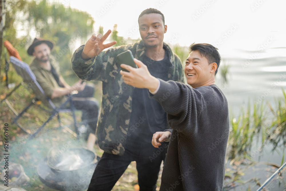 Smiling multiracial male friends taking selfie on smartphone during resting in nature. Men fishing a