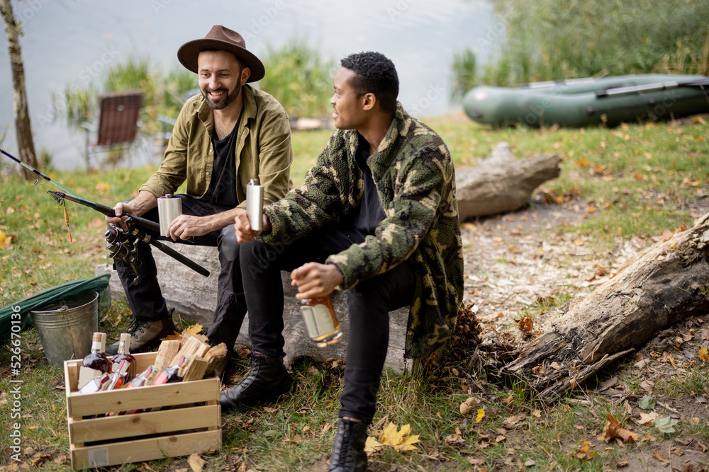 Two male friends sitting together, drinking some alcohol from flasks while fishing near lake. Caucas