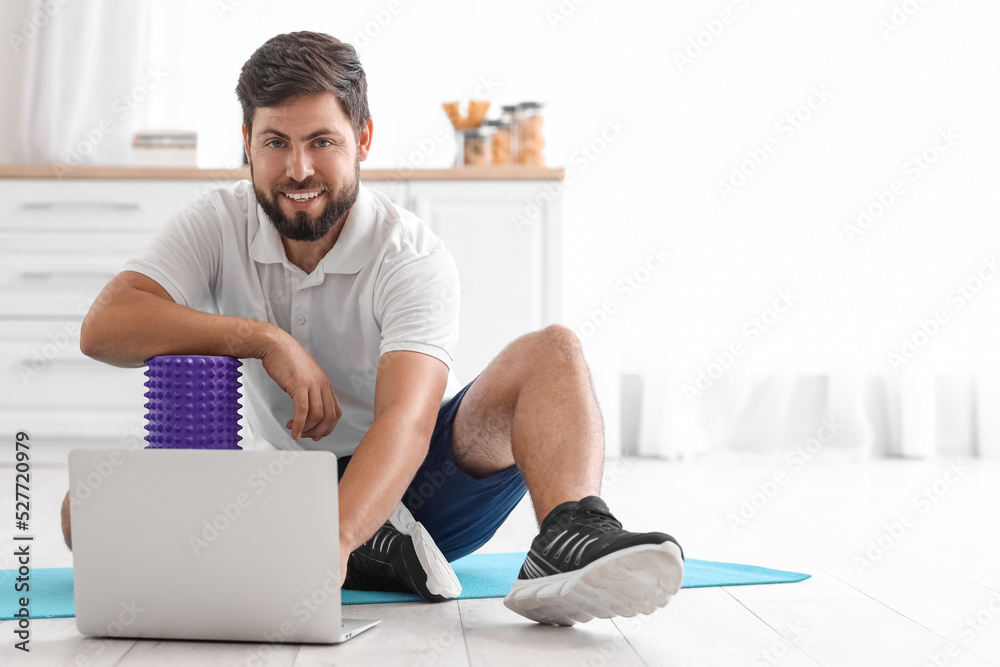 Young man with foam roller using laptop in kitchen