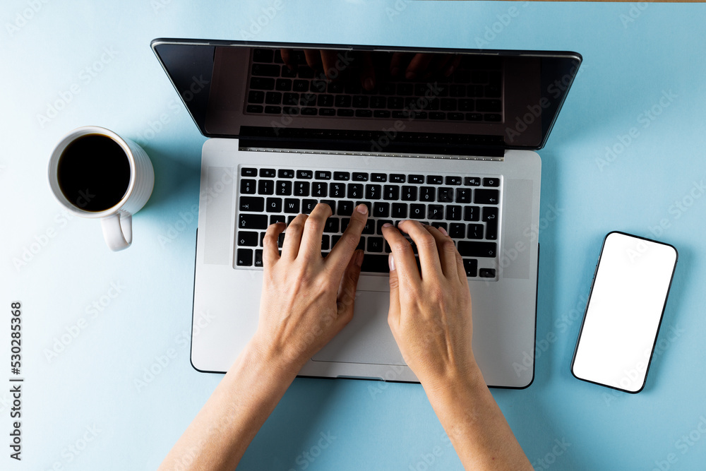 Composition of cup of coffee and hands using laptop on blue background