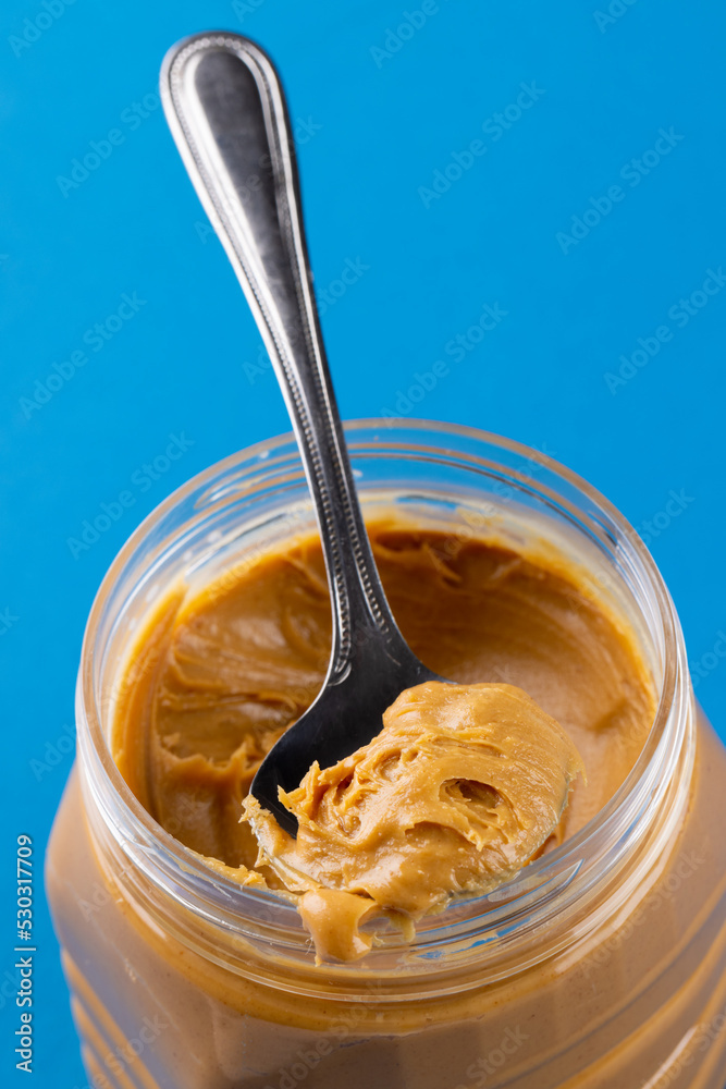 Vertical image of spoon and jar with peanut butter on blue surface