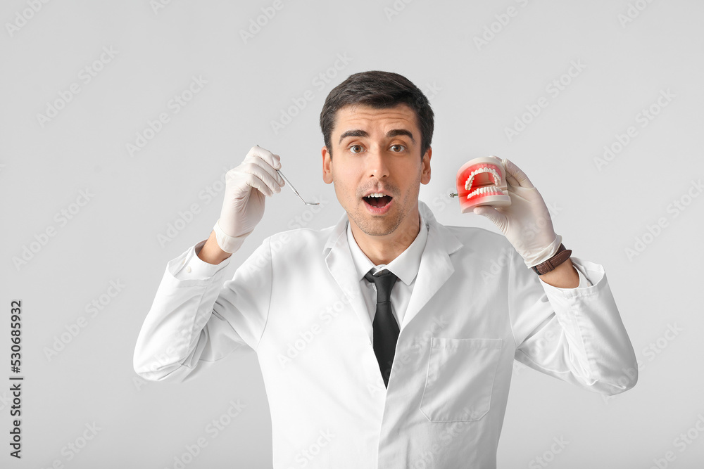 Male dentist with plastic jaw model and mirror on light background