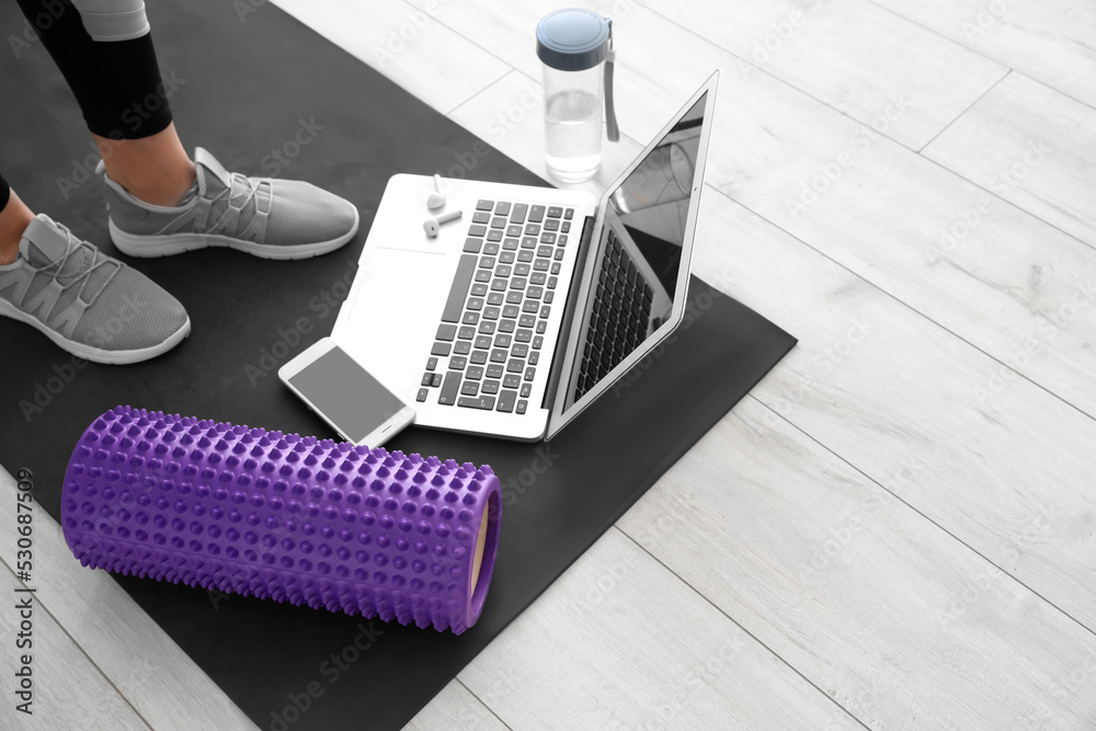 Woman with foam roller, mobile phone, laptop and bottle of water on mat at home