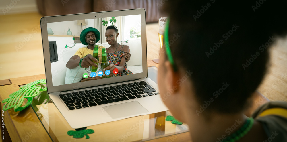 Mixed race woman making st patricks day video call to happy couple holding beers on laptop at home