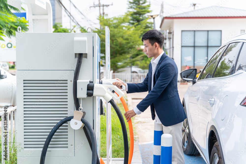 Asian man with EV Car or Electric vehicle at charging station with the power cable supply plugged on