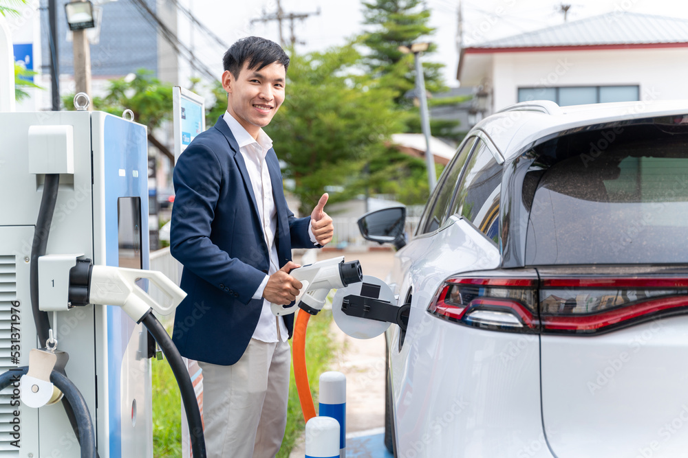 Asian man with electric car inserts the electrical connector to charge the batteries. Concept of gre