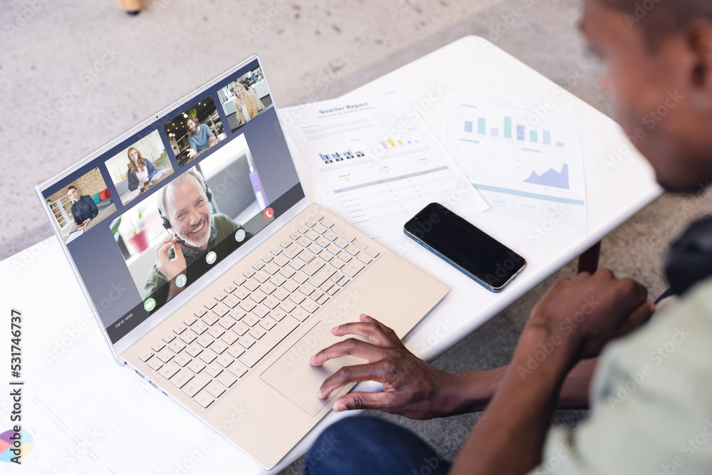 Multiracial man with cellphone and documents on desk discussing with coworkers on video call