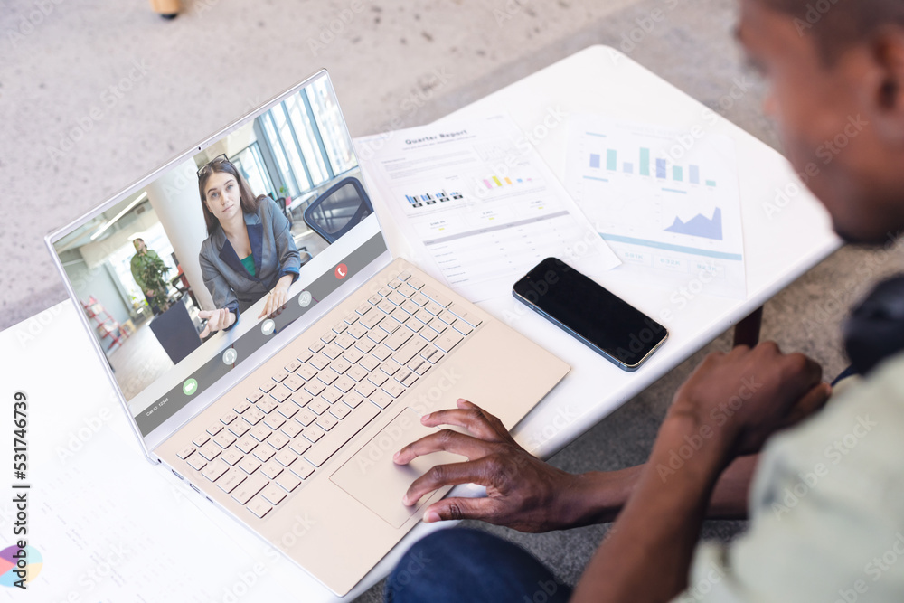 Diverse businessman with papers and phone on desk discussing with female coworkers on video call