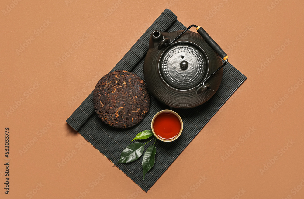 Bamboo mat with dry pressed puer tea, cup and teapot on brown background