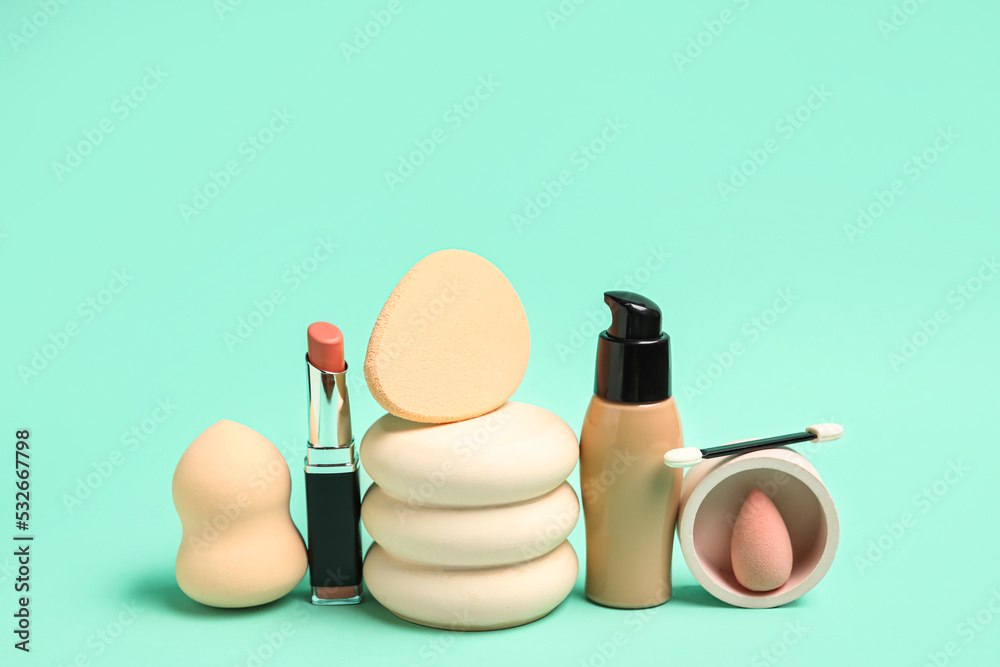 Makeup sponges with brush, lipstick, foundation and decor on green background