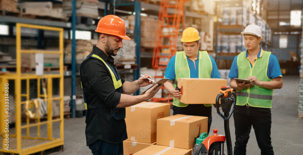 Warehouse manager wearing helmet pointing towards shelf using digital tablet and notepad in warehous