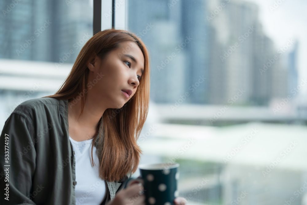 独自一人在家，想着孤独或悲伤的年轻亚洲女性坐在窗户边向外看